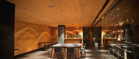 A modern interior with a wooden ceiling, recessed lighting, a central dining area with tables and chairs, and a kitchen area with shelves and appliances.