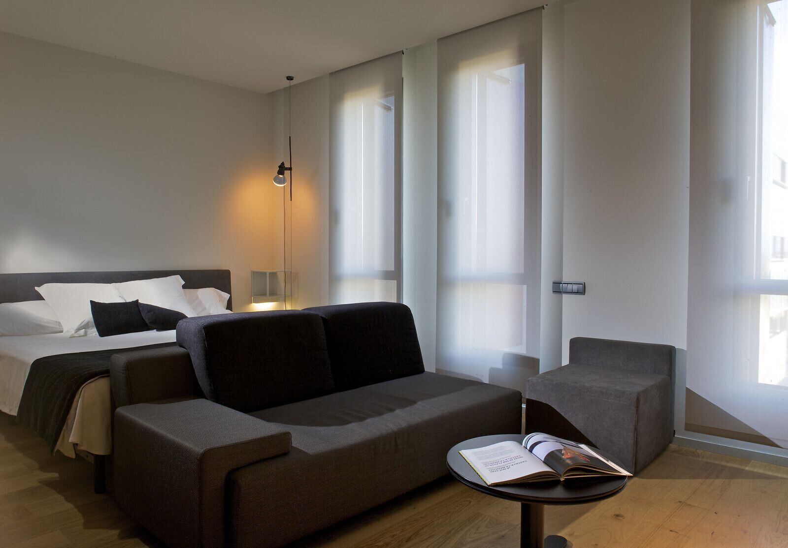 A modern hotel room with a bed, a sofa, a small table with a book, and large windows with sheer curtains.