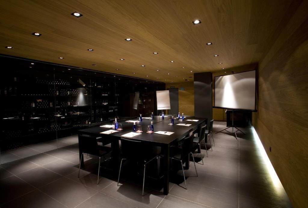 Un salón de conferencias con una larga mesa, sillas, una pantalla de proyector y una estantería para vinos al fondo.