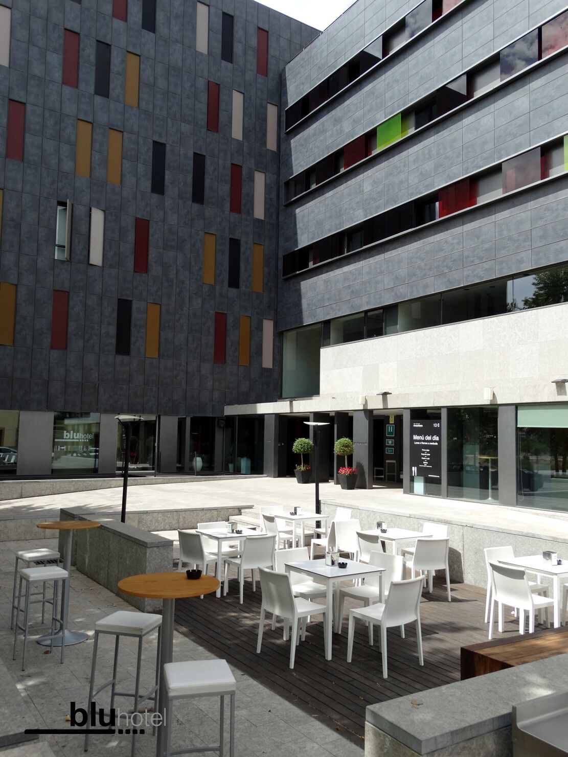 An outdoor seating area with white tables and chairs, a modern building with colorful window patterns, and a sign for 'blu hotel'.