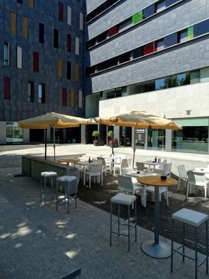 An outdoor seating area with tables and chairs, umbrellas, and a modern building with colorful windows in the background. - Blu Hotel Almansa (Almansa)