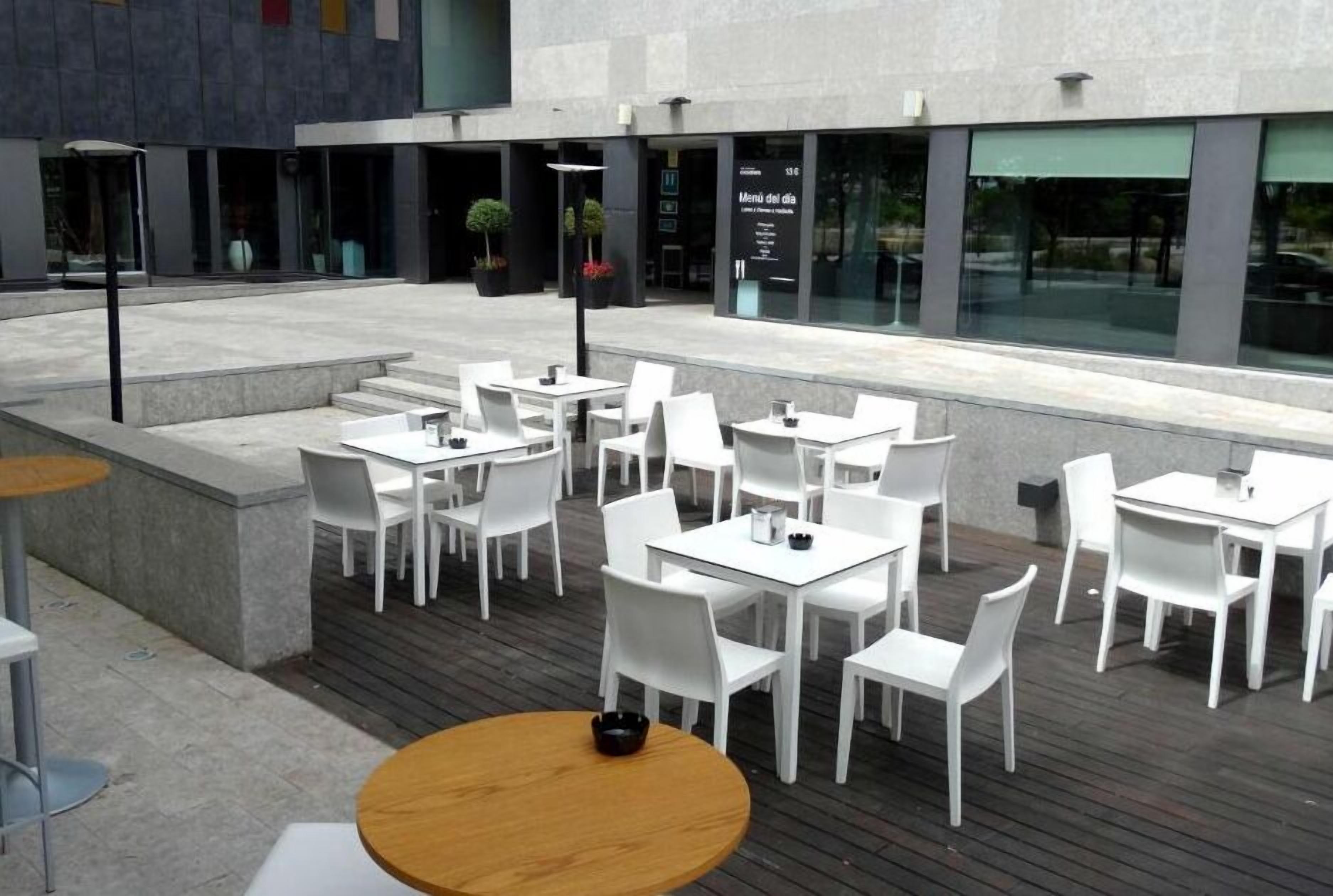 Un área de estar al aire libre con mesas y sillas blancas, una mesa de madera en primer plano y un edificio con un cartel de menú al fondo.