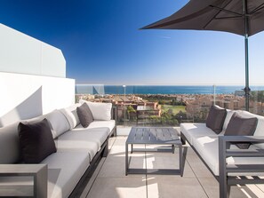 Un balcon avec un canapé blanc, une table en métal et un grand parasol. Le balcon offre une vue sur un paysage urbain côtier, avec des bâtiments et la mer.