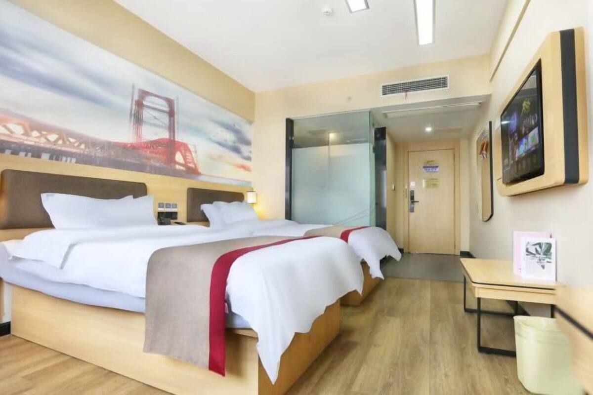 A hotel room with a bed, a desk, a chair, a trash bin, a painting, and a door.
