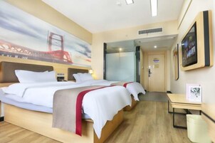 A hotel room with a bed, a desk, a chair, a trash bin, a painting, and a door.