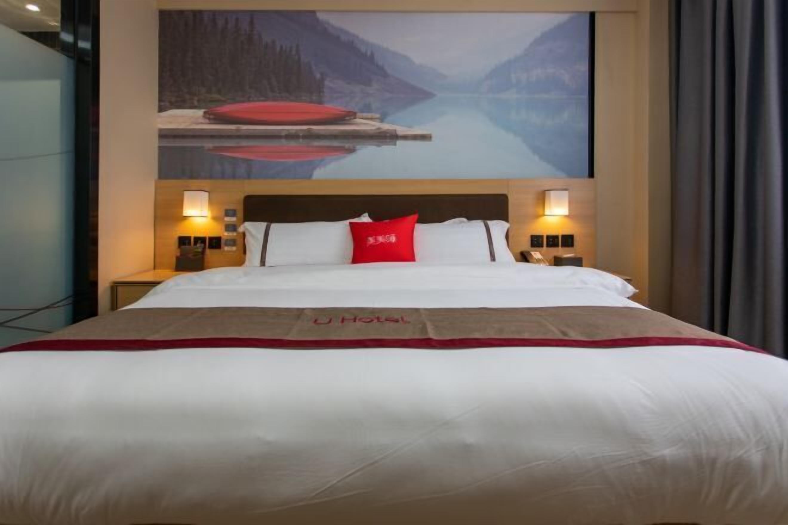Habitación de hotel con una cama grande, un almohadón rojo y un cuadro de un lago y montañas en la pared.