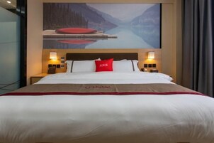 A hotel room with a large bed, a red pillow, and a painting of a lake and mountains on the wall.