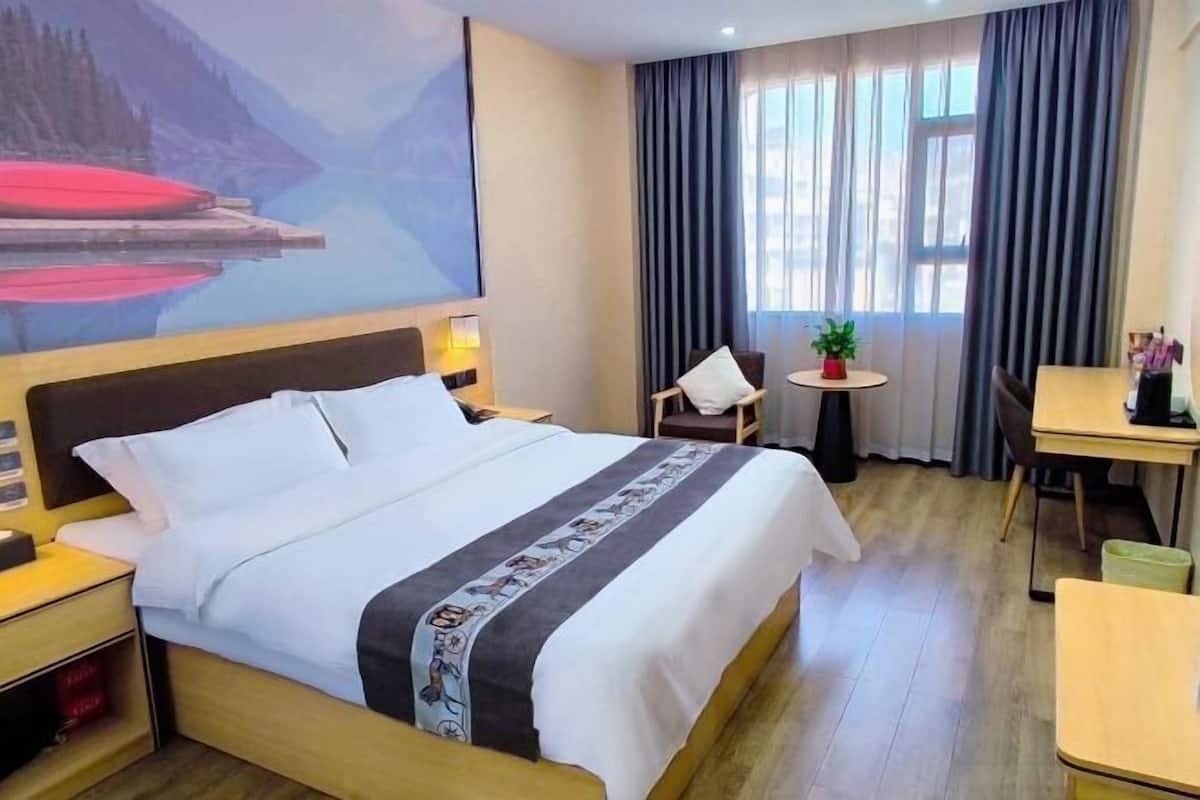 A hotel room with a bed, a desk, a chair, a small table with a plant, and a painting of a mountain landscape.