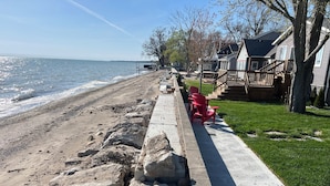 Une promenade en bord de mer, agrémentée d’une barrière en béton, de chaises rouges et d’une zone herbeuse menant à une maison dotée d’une terrasse.