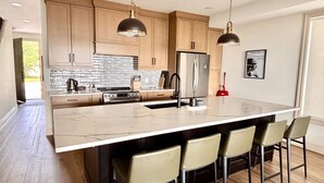 Une cuisine moderne avec des meubles en bois, un réfrigérateur en acier inoxydable, un grand îlot central doté de tabourets et un éclairage suspendu.