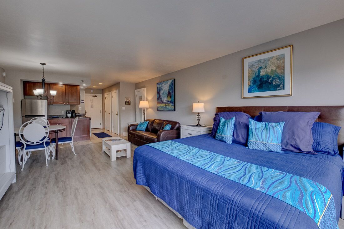 A hotel room with a large bed, a sitting area with a sofa and a small table, a kitchenette, and a dining area with a round table and chairs.