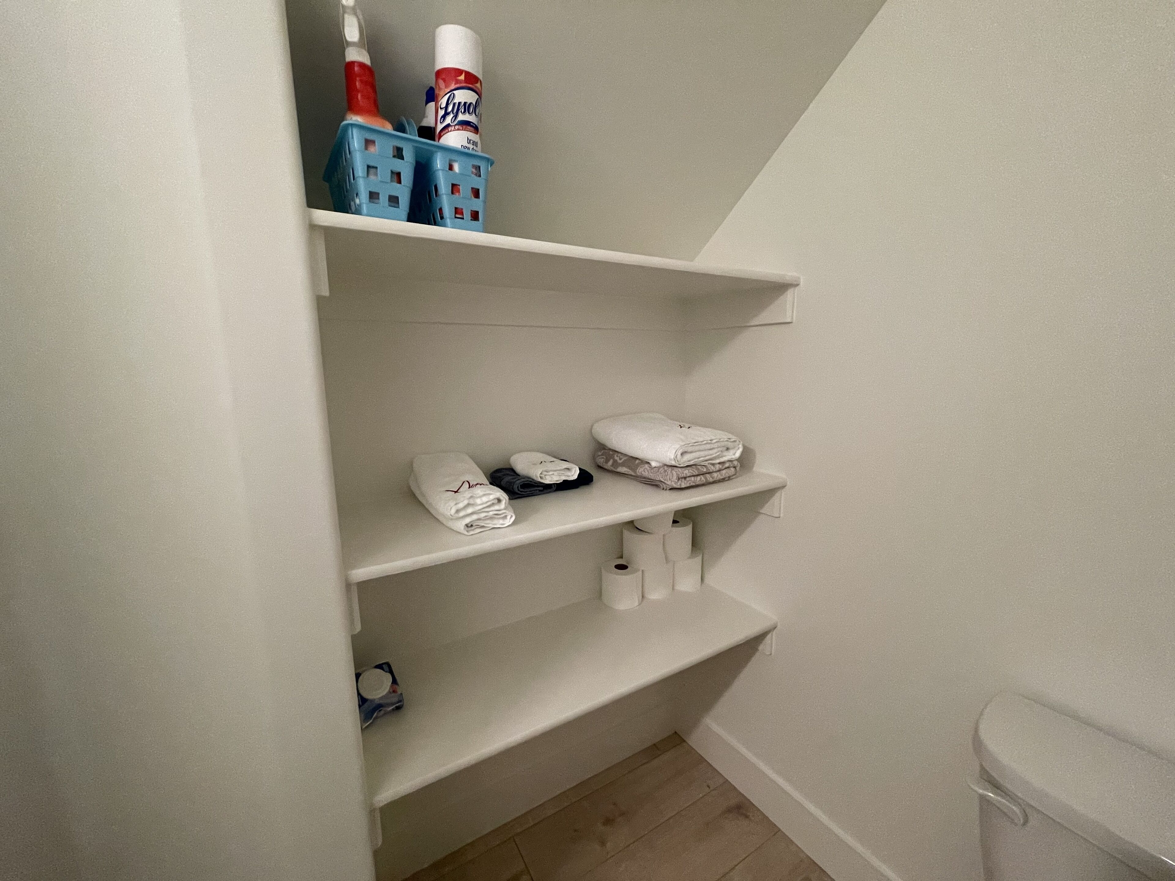 A bathroom with white shelves, a toilet, and a blue laundry basket containing toiletries.