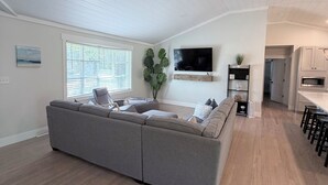 A modern living room with a sectional sofa, a flat-screen TV, and a kitchen area with bar stools. - Coastal Cottage in Charming Old Mt Pleasant! (Mount Pleasant)
