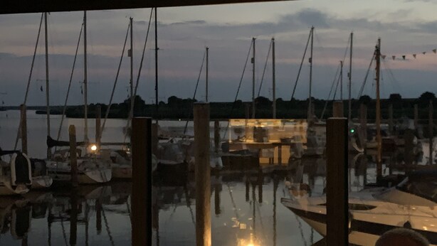 Un port de plaisance avec des bateaux amarrés à des quais en bois, une surface d’eau calme qui reflète les bateaux et un ciel dégagé en arrière-plan.