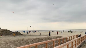 Une promenade en bois mène à une plage de sable où l’on peut voir des gens se promener et faire voler des cerfs-volants.