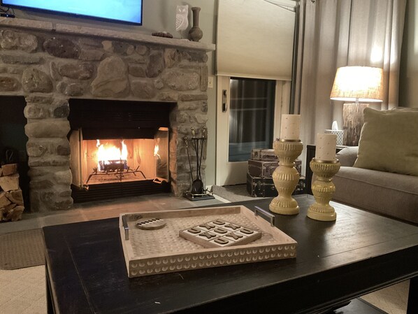 A living room with a stone fireplace, a flat-screen TV mounted above it, a coffee table with a tray, and a sofa with a lamp.
