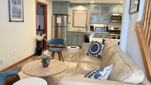 Un salon moderne avec un canapé en cuir beige, une table basse ronde et un fauteuil bleu. On aperçoit, au fond, une cuisine avec des meubles verts et des appareils électroménagers en acier inoxydable.