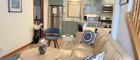 A modern living room with a beige leather sofa, a round coffee table, and a blue armchair. A kitchen with green cabinets and stainless steel appliances is visible in the background.
