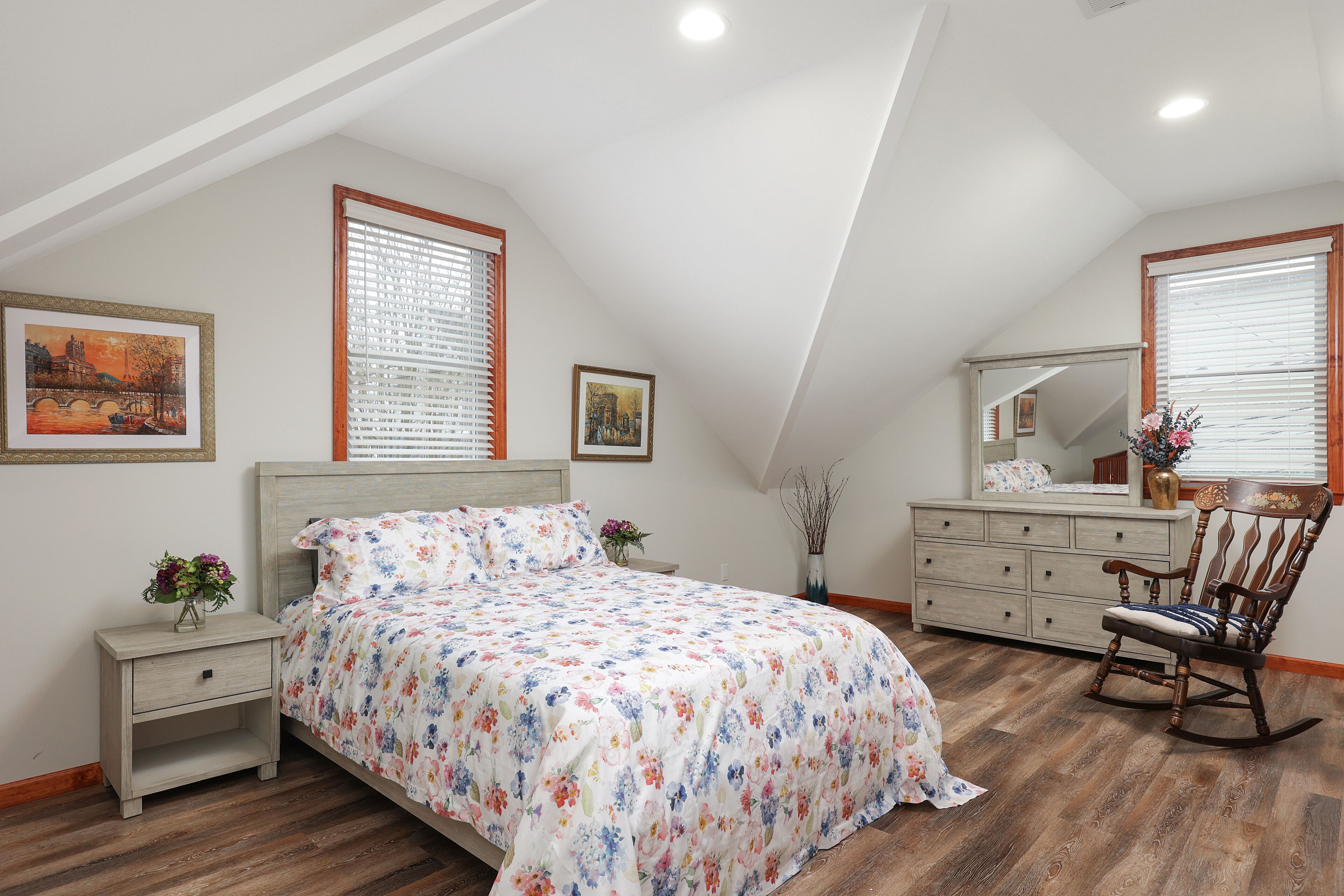 A bedroom with a bed, nightstand, dresser, rocking chair, and two windows with blinds.