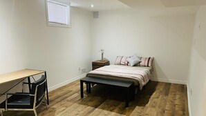 Un dormitorio con piso de madera, cama con ropa de cama estampada, escritorio con silla y mesita de noche con lámpara.