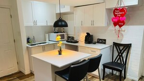 Una cocina moderna con armarios blancos, una isla central con sillas, una lámpara colgante negra y una decoración con forma de corazón que dice 'Happy Valentine's Day'.