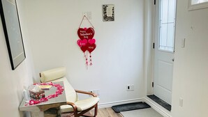 Una habitación con suelo de madera, una puerta y una pared blancos, un cuadro enmarcado, una decoración roja para el Día de San Valentín, una silla, una mesita con una caja de pañuelos y una botella, y un felpudo.