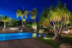 Un espace piscine nocturne doté d’une terrasse en bois, de palmiers et de sièges d’extérieur.