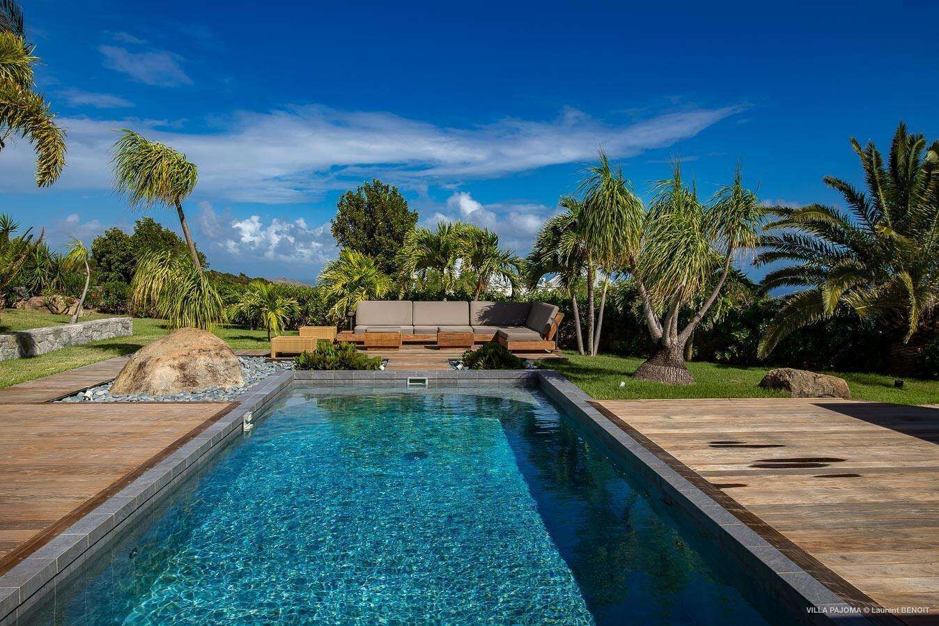 Une piscine avec une terrasse en bois, un espace détente comprenant un canapé et des fauteuils, le tout entouré de palmiers et de verdure.