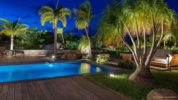Ein Poolbereich bei Nacht mit Holzterrasse, Palmen und Gartenmöbeln.
