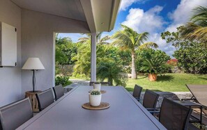 Un espace extérieur abrité, aménagé pour les repas, avec une table et des chaises, entouré d’une végétation luxuriante et de palmiers.