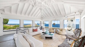 Amplia sala de estar con techo alto, ventanas grandes con vistas al océano, un conjunto de sofás, una mesa de centro y un sillón colgante.