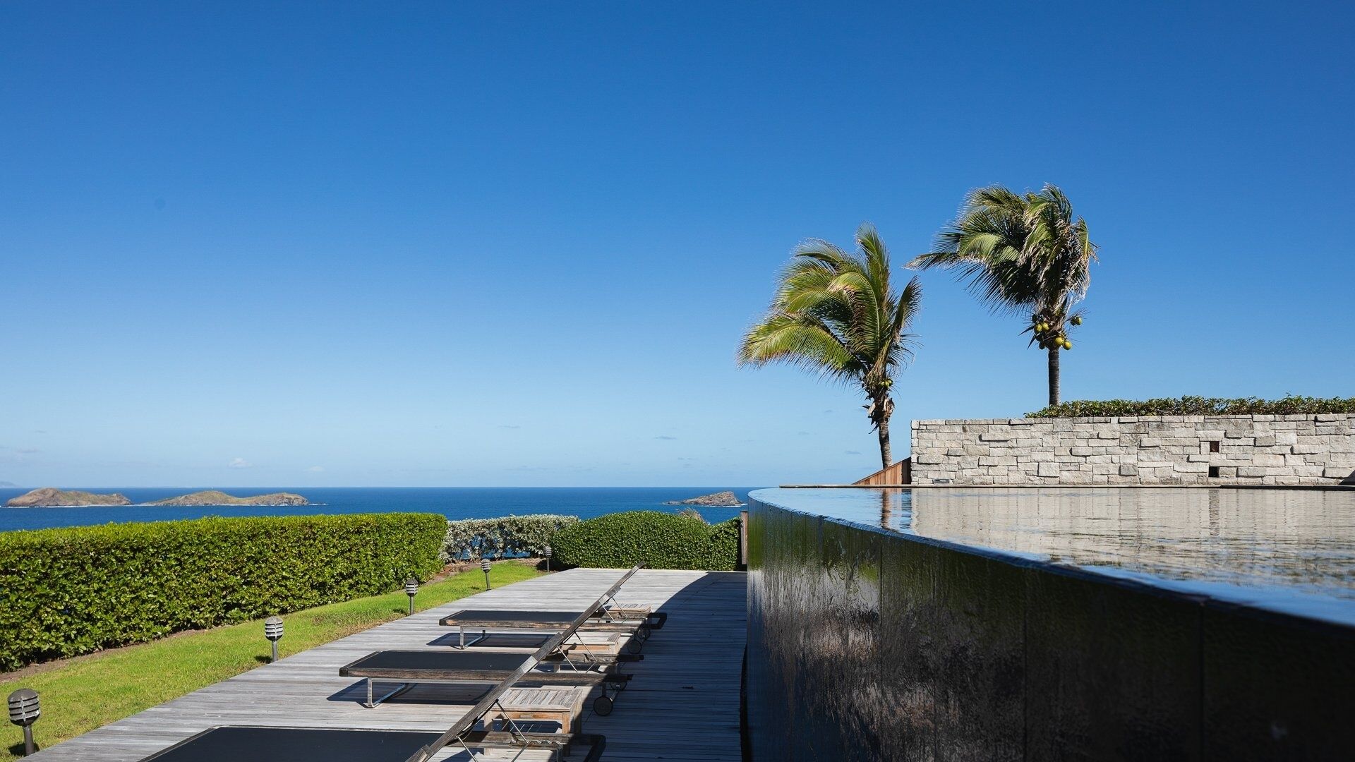 Un espace aménagé au bord de la piscine, avec des chaises longues, un mur en pierre et des palmiers offrant une vue sur l’océan.