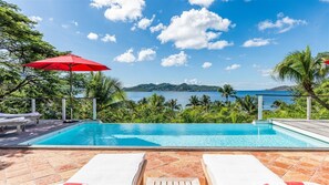 Uma piscina com espreguiçadeiras e um guarda-sol, rodeada por vegetação exuberante e vista para o mar.