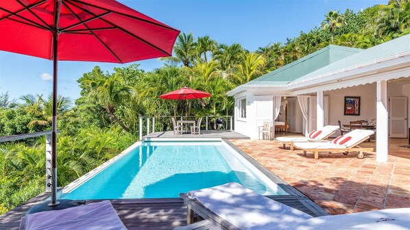 A pool area with a red umbrella, lounge chairs, and a view of a house with a covered patio. - Alizee d'Eden (1/2 Rooms) (Pointe Milou)