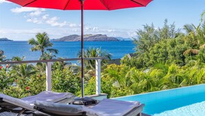 A poolside view with lounge chairs, a red umbrella, and a view of the ocean and mountains. - Alizee d'Eden (1/2 Rooms) (Pointe Milou)