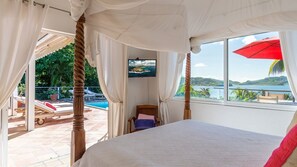 A bedroom with a large bed, a TV, and a view of a pool and greenery. - Alizee d'Eden (1/2 Rooms) (Pointe Milou)