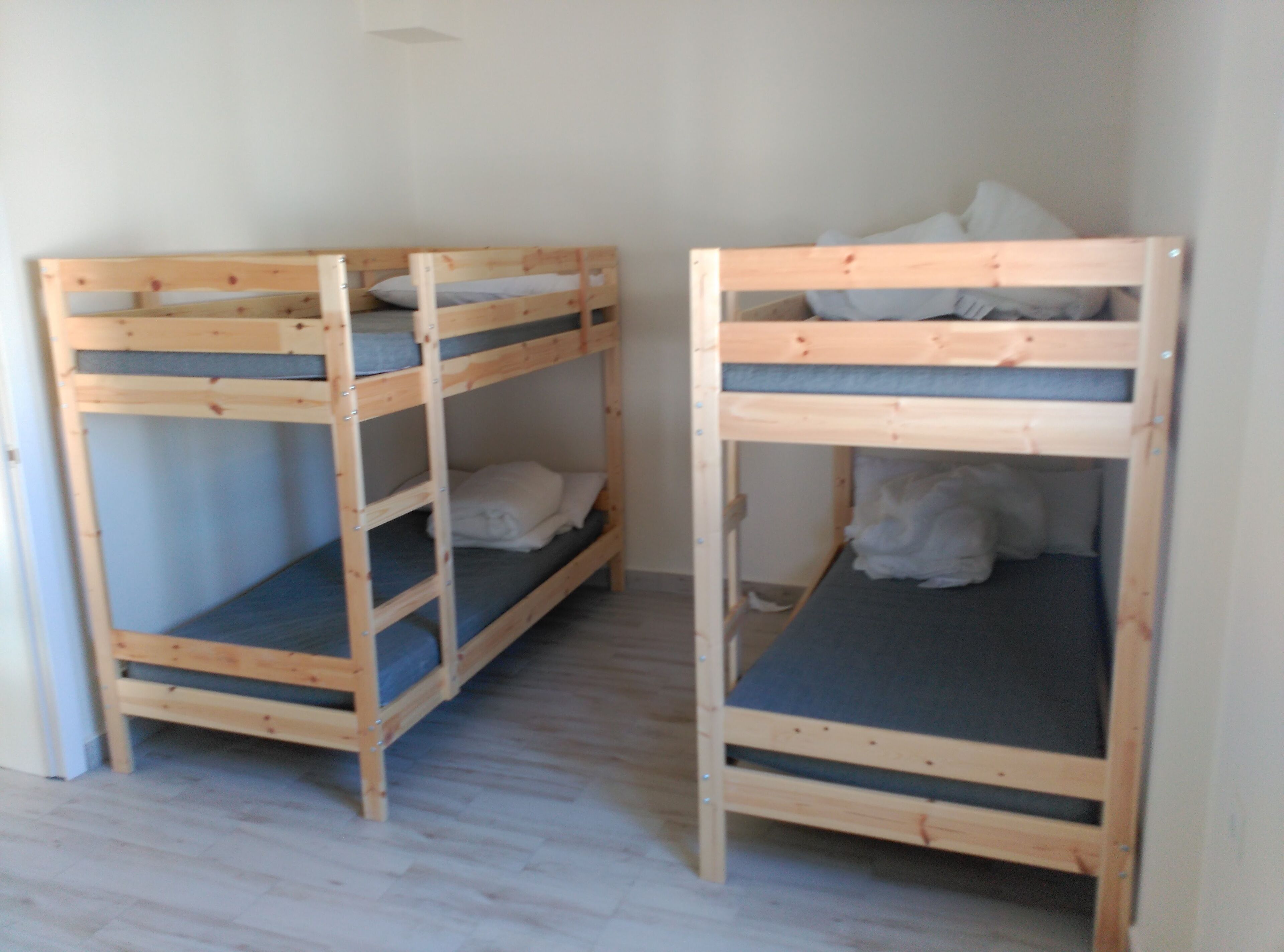 Dos literas con estructura de madera y ropa de cama gris, en una habitación con suelo de madera clara y paredes blancas.