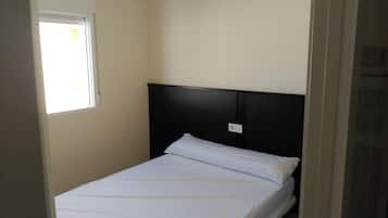 A small, single-bed room with a plain white bed, a black headboard, and a window with blinds.