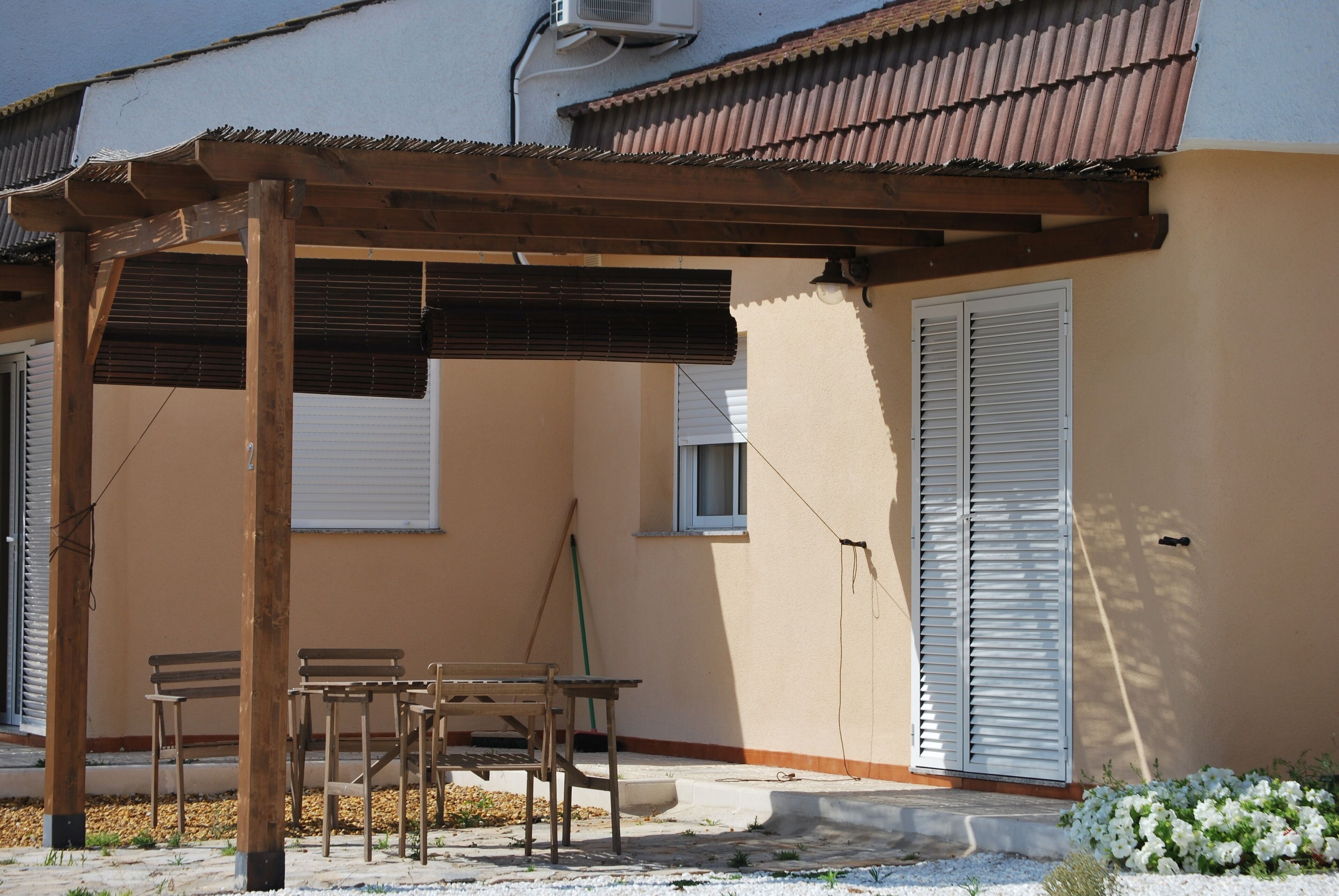 A covered outdoor seating area with wooden furniture, a pergola, and a building with a beige facade and white shutters.