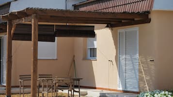 A covered outdoor seating area with wooden furniture, a pergola, and a building with a beige facade and white shutters.
