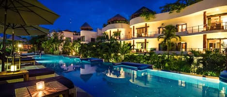 A resort pool area with lounge chairs and umbrellas, surrounded by lush greenery and buildings with thatched roofs.