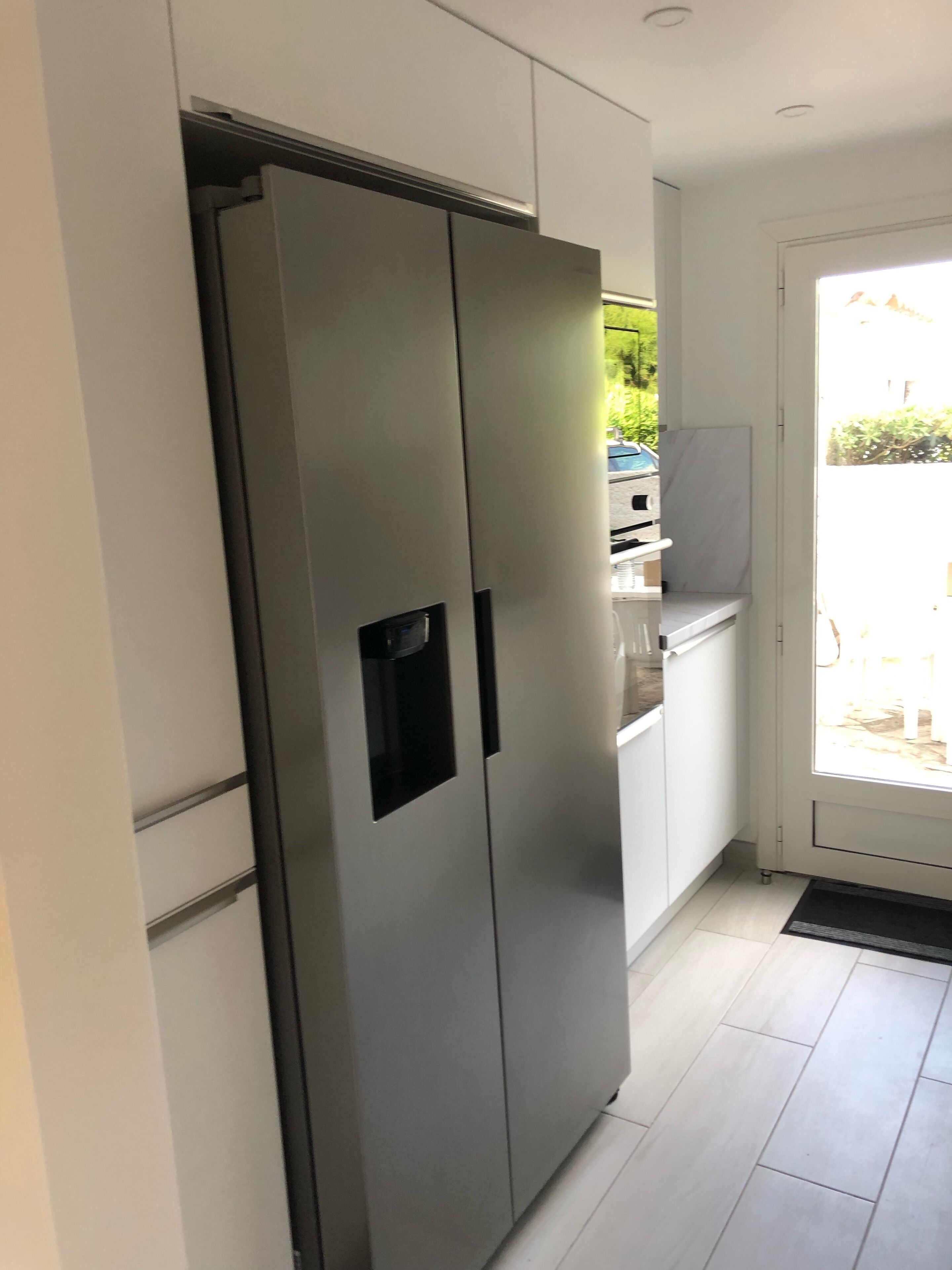 Een moderne keuken met een inbouwkoelkast van roestvrij staal, witte kastjes en uitzicht op de omgeving door een glazen deur.