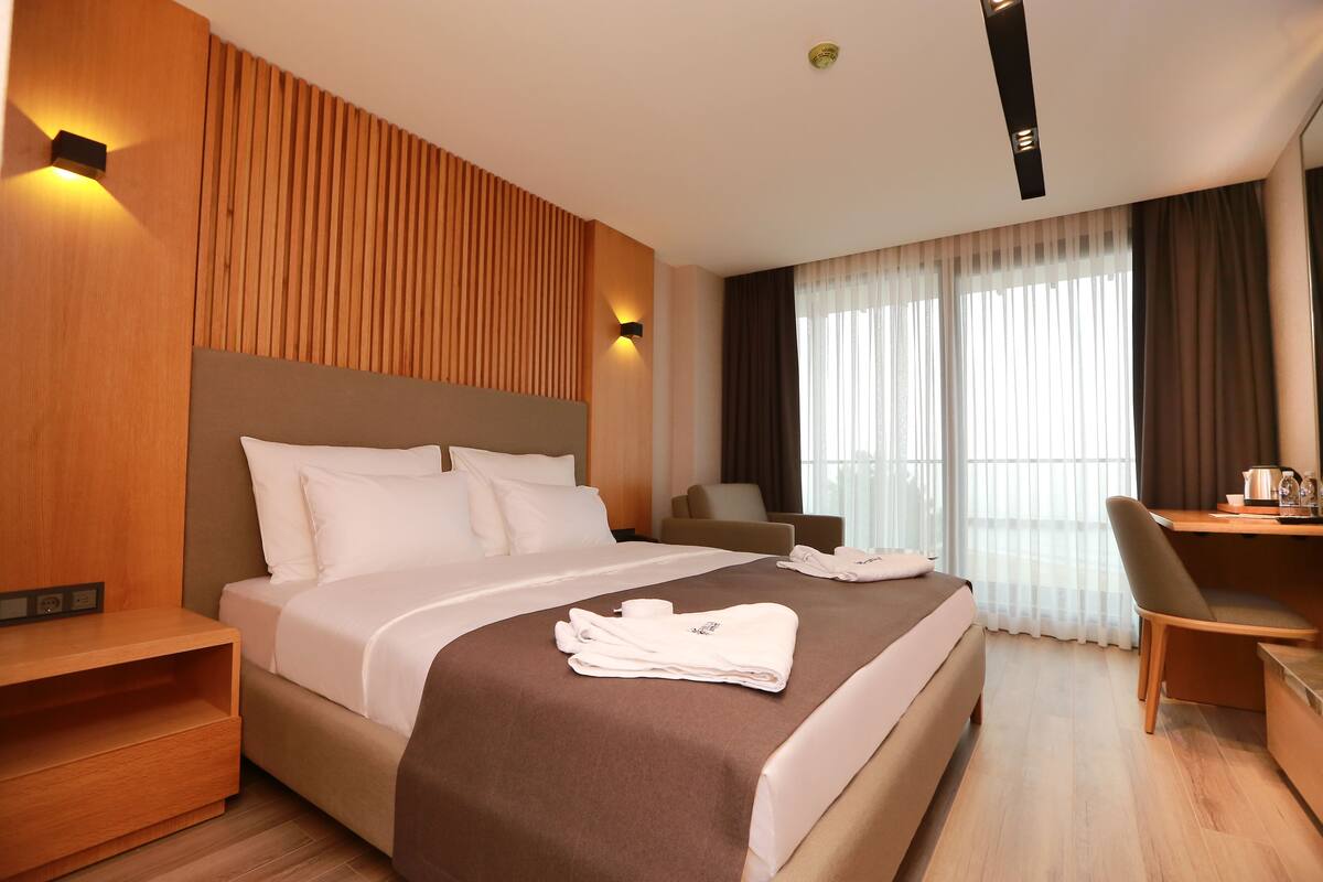 Quarto de hotel moderno com uma cama grande, uma escrivaninha e uma cadeira. Há duas luzes fixadas na parede e uma janela com cortinas.
