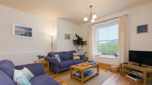 A living room with a sofa, a coffee table, a TV, and a window with curtains. - Chynance (Penzance)