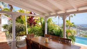 Un espace extérieur abrité, aménagé pour les repas, avec une table et des chaises, entouré d’une végétation tropicale luxuriante et offrant une vue sur une piscine et un paysage verdoyant.