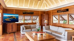 Un salon spacieux aux murs en bois, avec une grande télévision, deux canapés, une table basse et une vue sur la mer par les fenêtres.