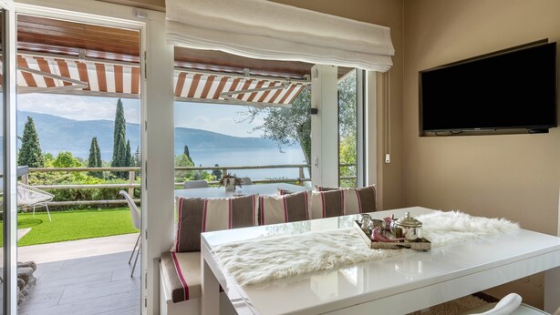 A room with a white table, a flat-screen TV mounted on the wall, and a view of a green lawn and mountains through large windows. - Apartment "Casa 50" with Garden & Shared Pool (Gargnano)