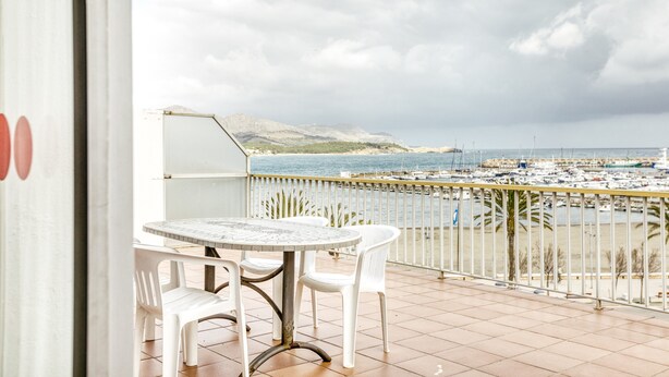 Un balcón con mesa y sillas, con vista a un puerto deportivo con barcos y una montaña distante.