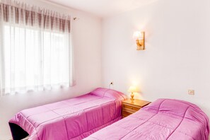 A bedroom with two beds, a bedside table, a lamp, and a window with sheer curtains.