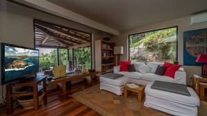 A modern living room with a large TV, wooden furniture, a sofa with red cushions, and a bookshelf. - Kuban (2/3 Rooms) Luxury villa (Colombier)